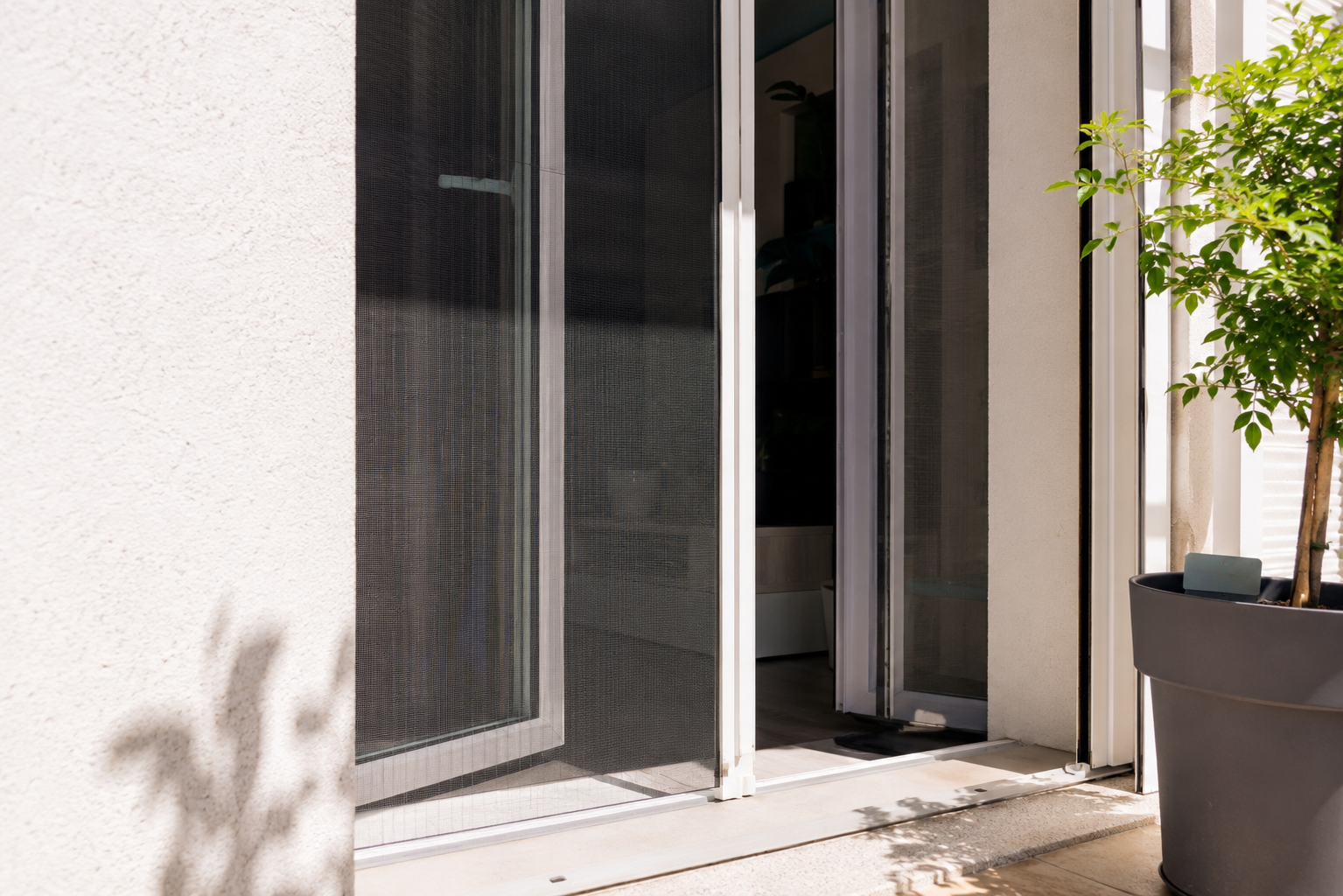 Porta scorrevole in vetro scuro aperta su patio, con piante verdi in fioriera e luce solare.